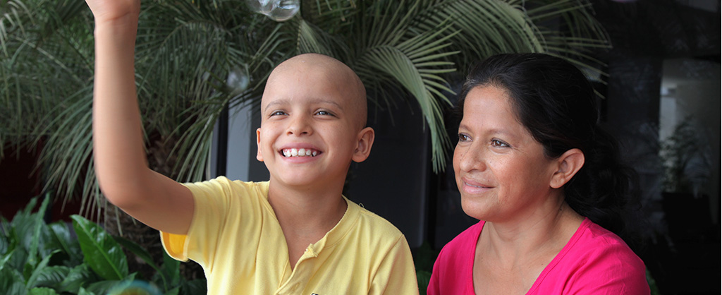 Niño calvo levantando su mano para saludar, mientras sonríe, al lado de su madre.