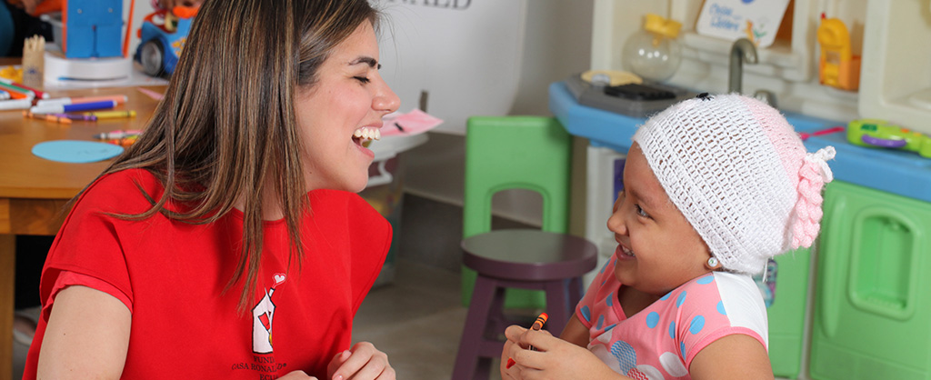 En una sala, hay una mujer, que tiene una camiseta roja con el logo de la Fundación Casa Ronald McDonald Ecuador, ríe al lado de a niña quien sostiene una crayola en su mano y dibuja.