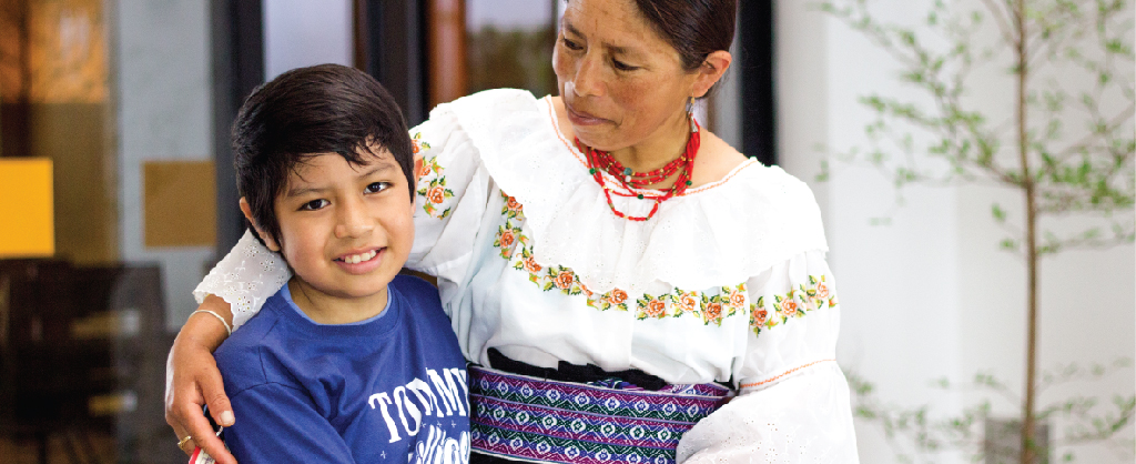 Una madre vistiendo un atuendo típico mexicano, abraza a su hijo quien sonríe.